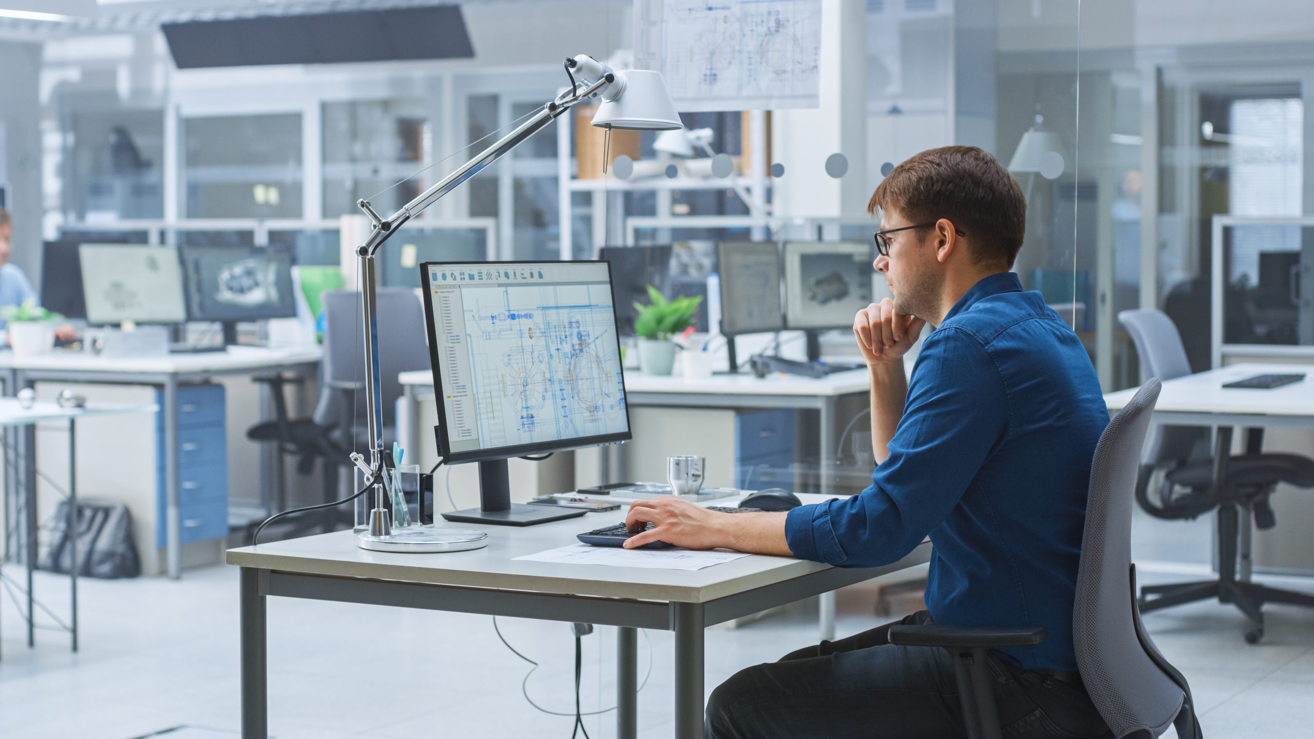 Over the Shoulder Shot of Engineer Working with CAD Software on Desktop Computer, Screen Shows Technical Drafts and Drawings. In the Background Engineering Facility Specialising on Industrial Design